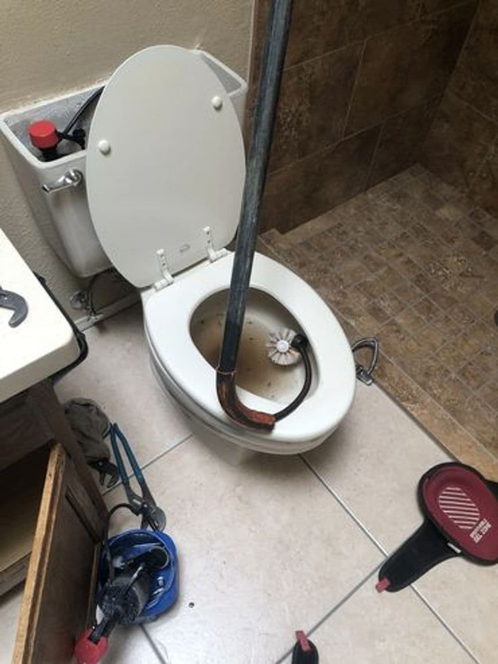 Broken toilet with a plumbing snake inserted, surrounded by tools and tile flooring.
