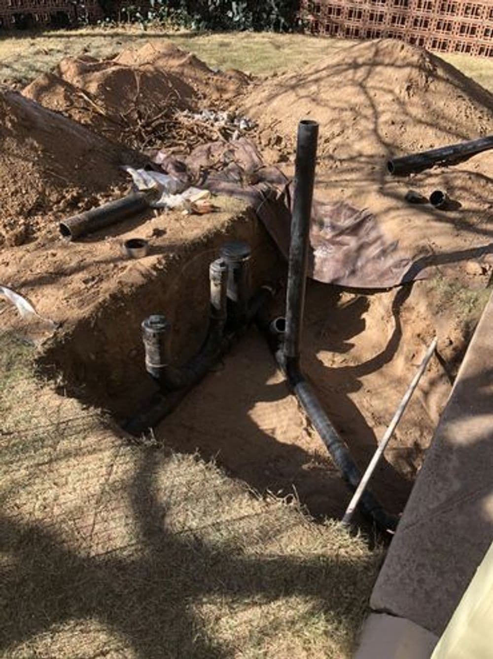 Excavated area in yard showing exposed black plumbing pipes and sandy soil.
